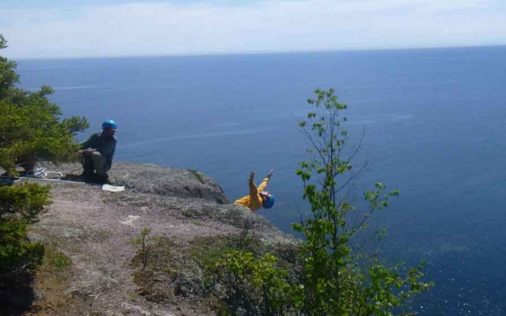 A person wearing safety gear is secured by ropes as they lean back from a cliff and throw their hands into the air. They are high above a blue body of water, and another person watches them from on top of the cliff. 